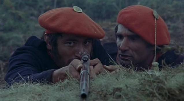 Fotografía de dos soldados detrás de una trinchera. Ambos llevan uniforme y gorra roja. El de la izquierda tiene una escopeta en las manos y parece que está a punto de disparar. El de la derecha le está hablando.