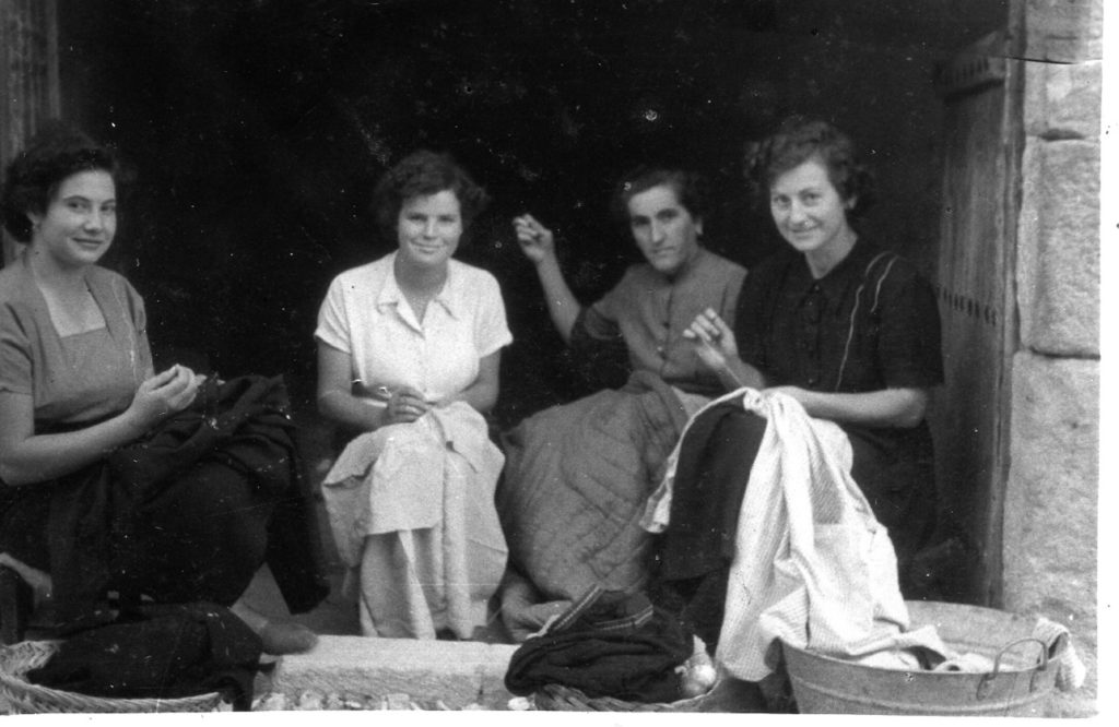 Imagen de cuatro mujeres cosiendo, sentadas en la entrada de una casa. La luz del sol las ilumina y el interior del edificio se ve oscuro. Las cuatro nos miran sonrientes.