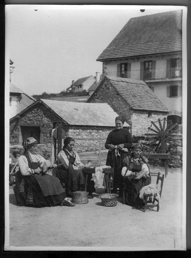 Fotografía de cuatro mujeres: tres sentadas en una silla y una de pie junto al txoatile, un instrumento de madera de dos piezas que se usa para hilar la lana de oveja latxa. Las cuatro mujeres están en la calle y detrás podemos apreciar tres casas del pueblo. 