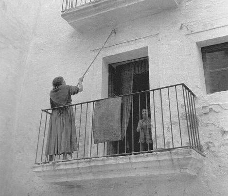 Imagen de un balcón sacada desde abajo. En el balcón hay una mujer encalando la fachada de la casa con un palo largo. Dentro de la casa vemos a un niño que mira hacia la cámara.