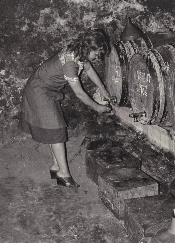 Fotografía en blanco y negro de una mujer ligeramente agachada, sirviéndose el pacharán del grifo de un barril.