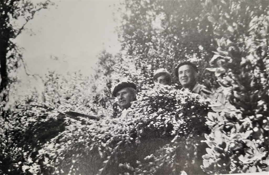 Fotografía de 3 hombres mirándonos sonrientes escondidos detrás de unos arbustos. Uno de ellos parece que esté apuntando con una escopeta a algún animal a la izquierda de la imagen.